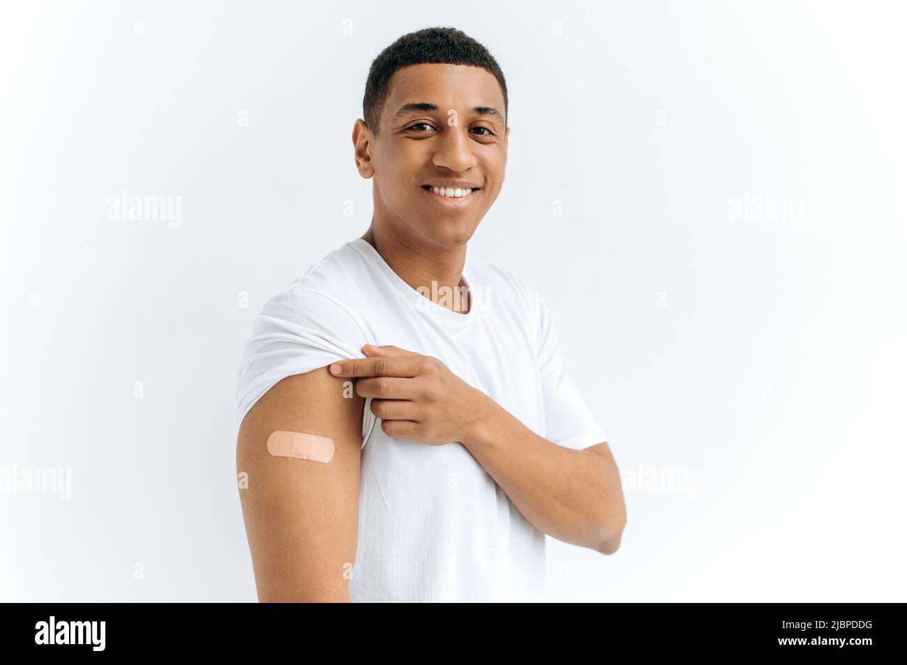 Mixed Race Happy Guy steht auf isoliertem weißem Hintergrund mit einem Gips auf seiner Schulter, erhielt einen Impfstoff gegen verschiedene Krankheiten, schaut auf die Kamera, lächelt. Gesundheits- und Immunitätskonzept Stockfoto