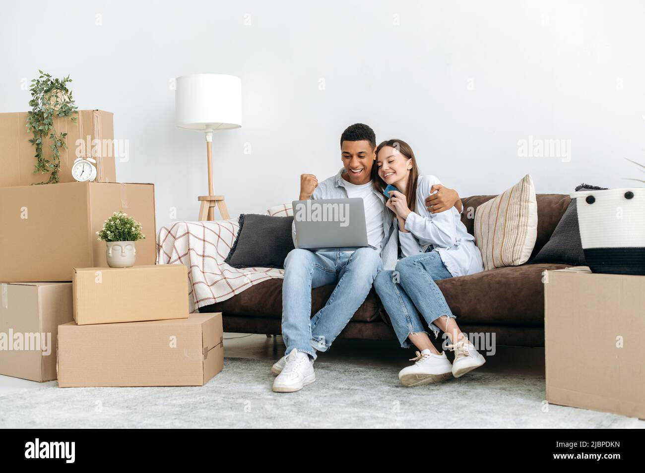 Immobilienkonzept. Ein glückliches gemischtes Familienpaar sitzt auf einem Sofa zwischen Pappkartons im Wohnzimmer ihres neuen Zuhauses, nutzt Laptop und Kreditkarte für Online-Shopping und kauft Dinge für zu Hause Stockfoto