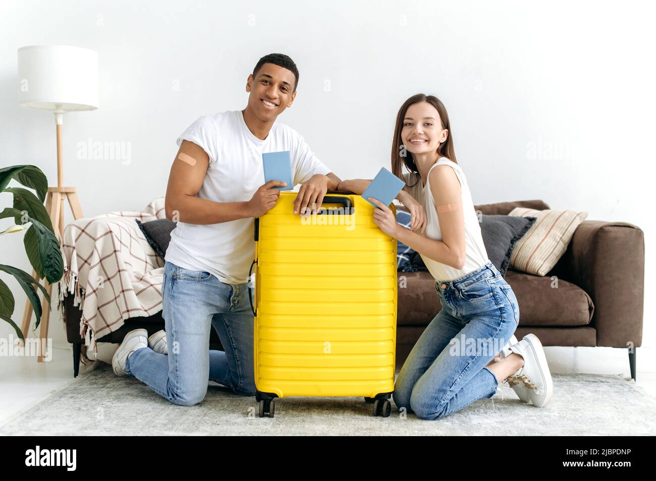 Geimpfte Typen und Mädchen verschiedener Nationalitäten, mit ihren Pässen, sitzen auf dem Boden im Wohnzimmer neben dem Koffer mit Dingen, gehen nach der Pandemie in den Urlaub, lächeln glücklich Stockfoto