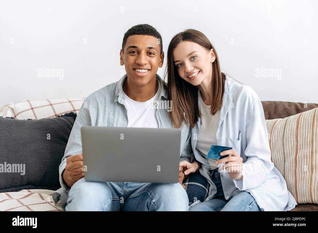 Online-Shopping. Glückliches, positives gemischtes Familienpaar, das auf einem Sofa im gemütlichen Wohnzimmer sitzt, Laptop und Kreditkarte zum Online-Shopping nutzt, Dinge für zu Hause kauft, Lieferung nach Hause bestellt, lächelt Stockfoto