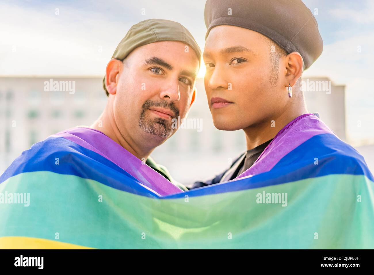Ein Paar von verliebten schwulen Männern umarmt sich gegenseitig, eingewickelt in eine schwule Flagge, in einem Park bei Sonnenuntergang, mit Reflexen der Sonne. Konzept von Vielfalt, Stolz, Stockfoto