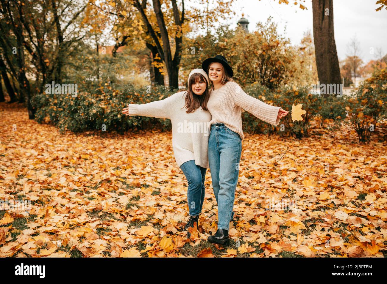 Zwei junge Freundinnen haben Spaß im Park, in dem sie mit ihren Armen umeinander und ausgestreckten Armen posieren. Sie sind in blauen Jeans und gekleidet Stockfoto