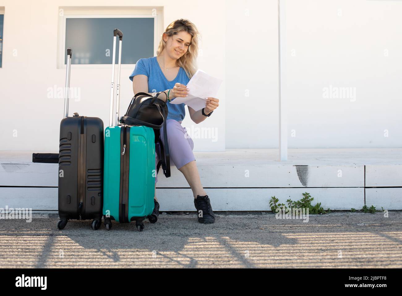 Blonde, frohe Frau mit Gepäck, das in der Nähe des Bahnhofs sitzt und auf den Transport wartet. Starten Sie ein neues Leben im Ausland. Abfertigung Stockfoto