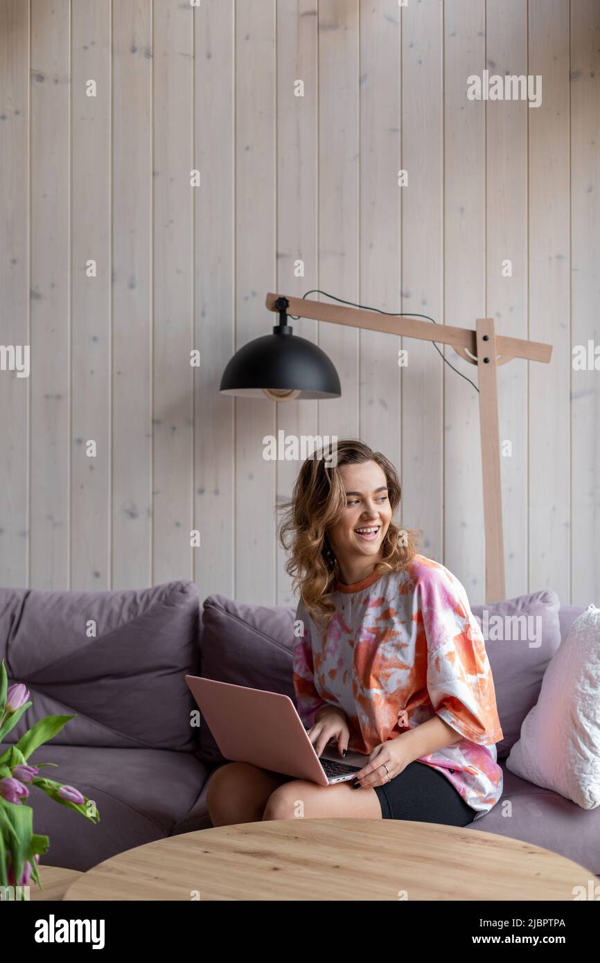 Lachende, charmante, braunhaarige Frau mit langen Haaren in bequemer Heimkleidung sitzt zu Hause im Internet auf einem gemütlichen Sofa an der Holzwand Stockfoto