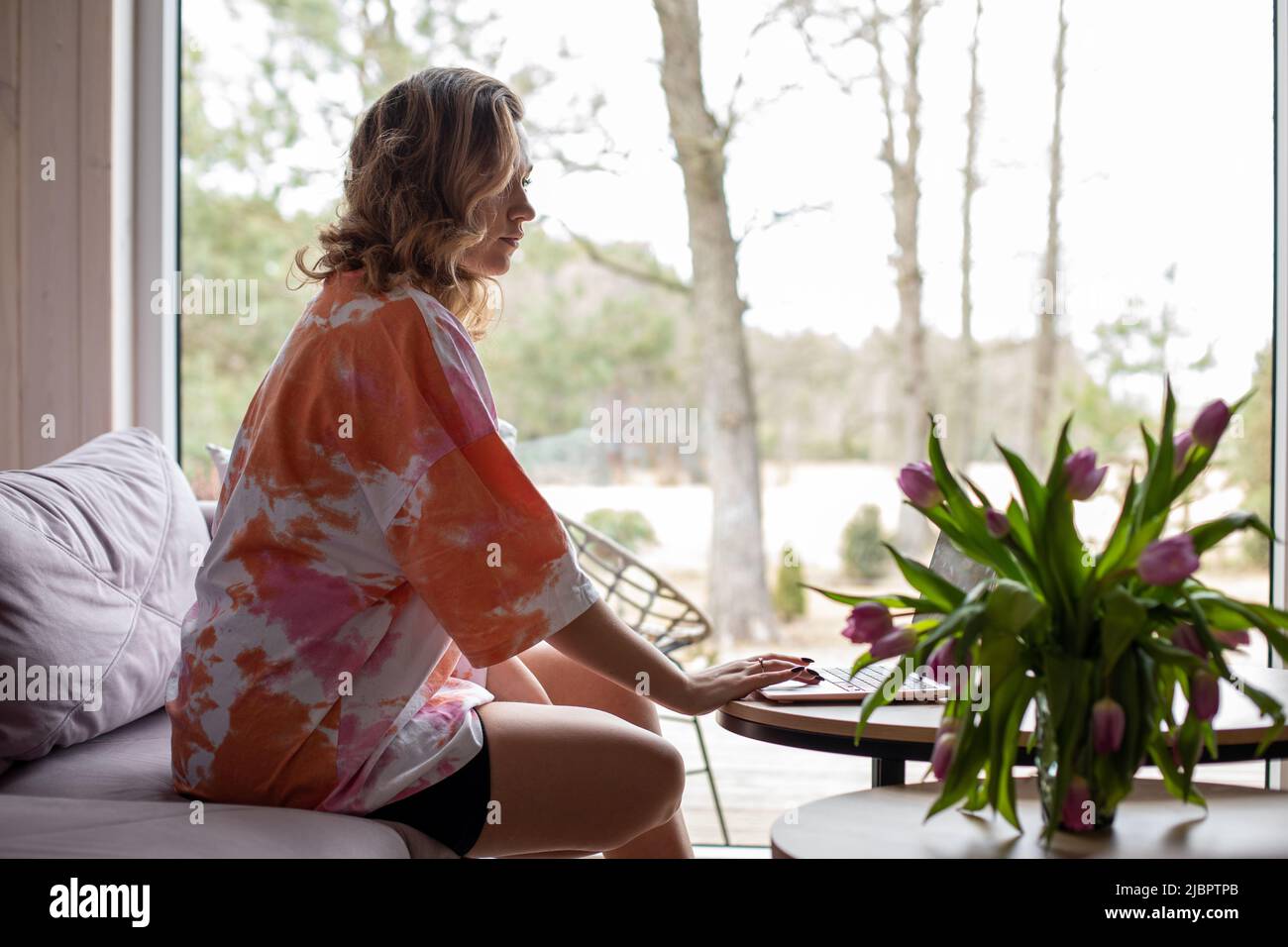 Junge Frau mit langen lockigen Haaren in lockerer Hauskleidung arbeitet zu Hause online auf einem gemütlichen Sofa, gegenüber dem Panoramafenster sitzend. Geschäftsfrauen. Modern Stockfoto