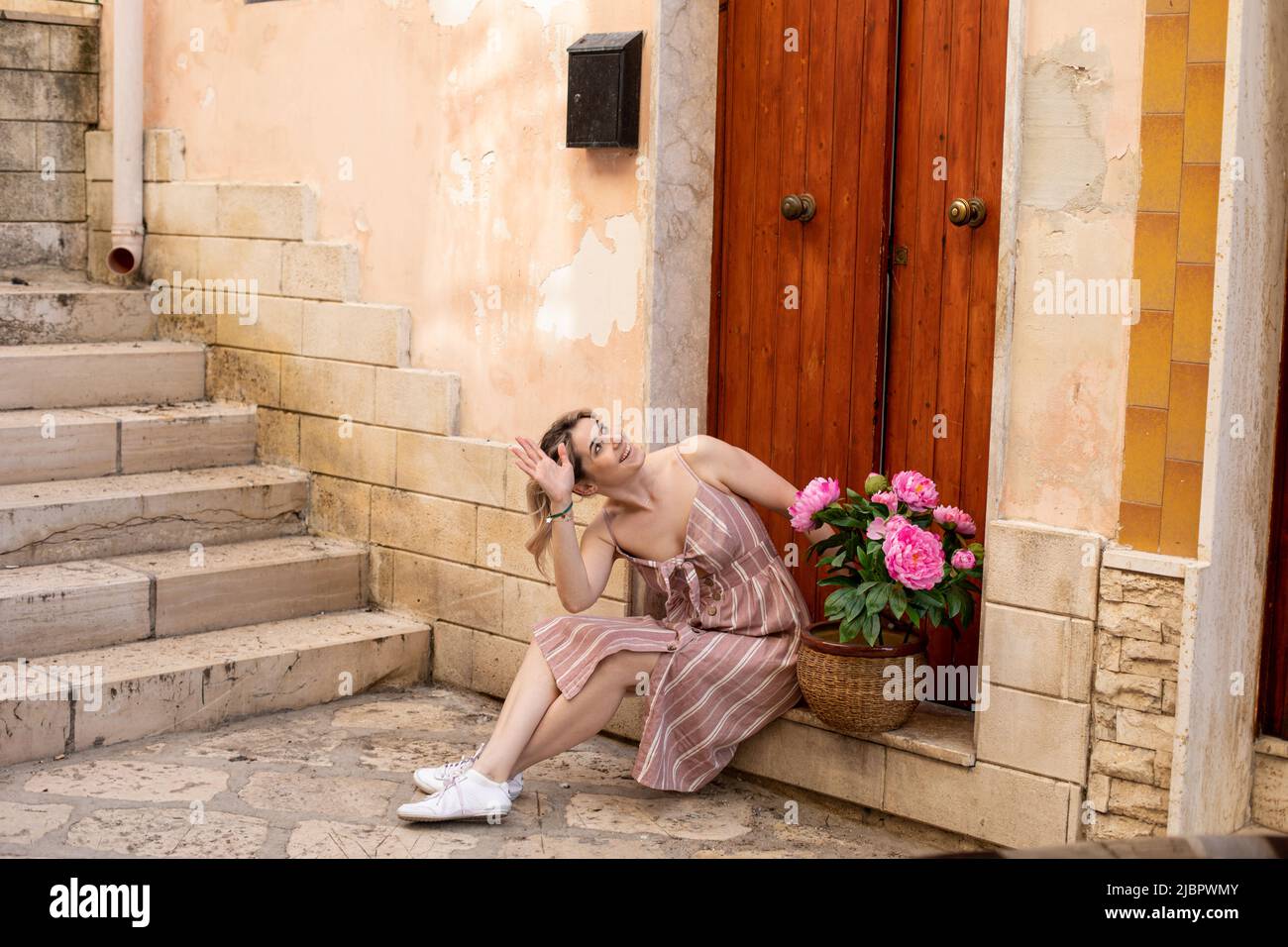 Junge fröhliche Frau in rosa gestreiftem Kleid, sitzt auf einer Stufe neben der roten Holztür, schaut nach oben, hebt die Hand, grüßt. Stockfoto