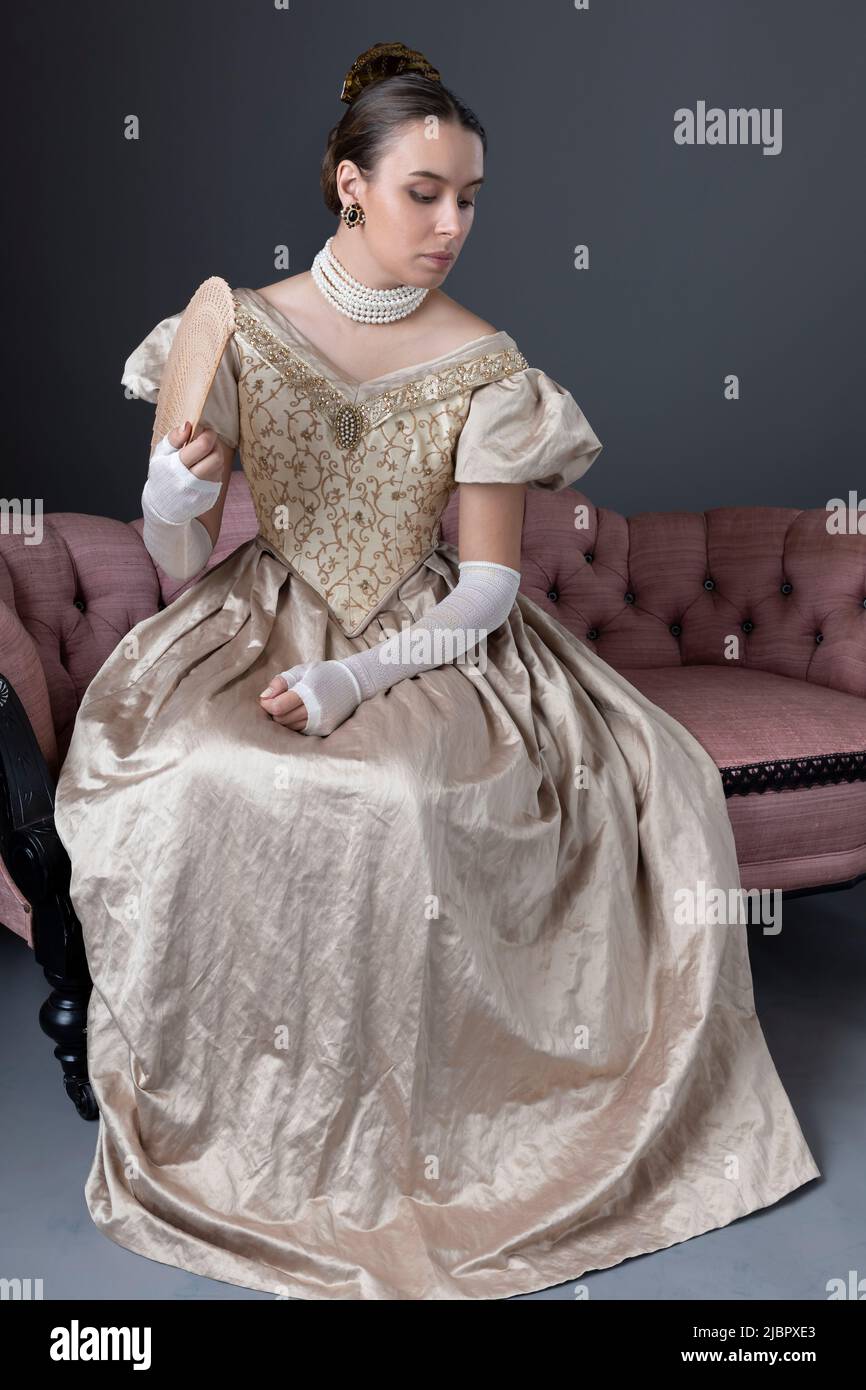 Eine viktorianische Frau, die ein goldenes Ballkleid trägt und auf einem rosa Sofa sitzt Stockfoto