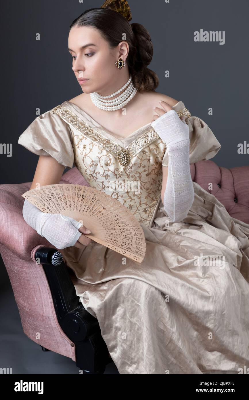 Eine viktorianische Frau, die ein goldenes Ballkleid trägt und auf einem rosa Sofa sitzt Stockfoto