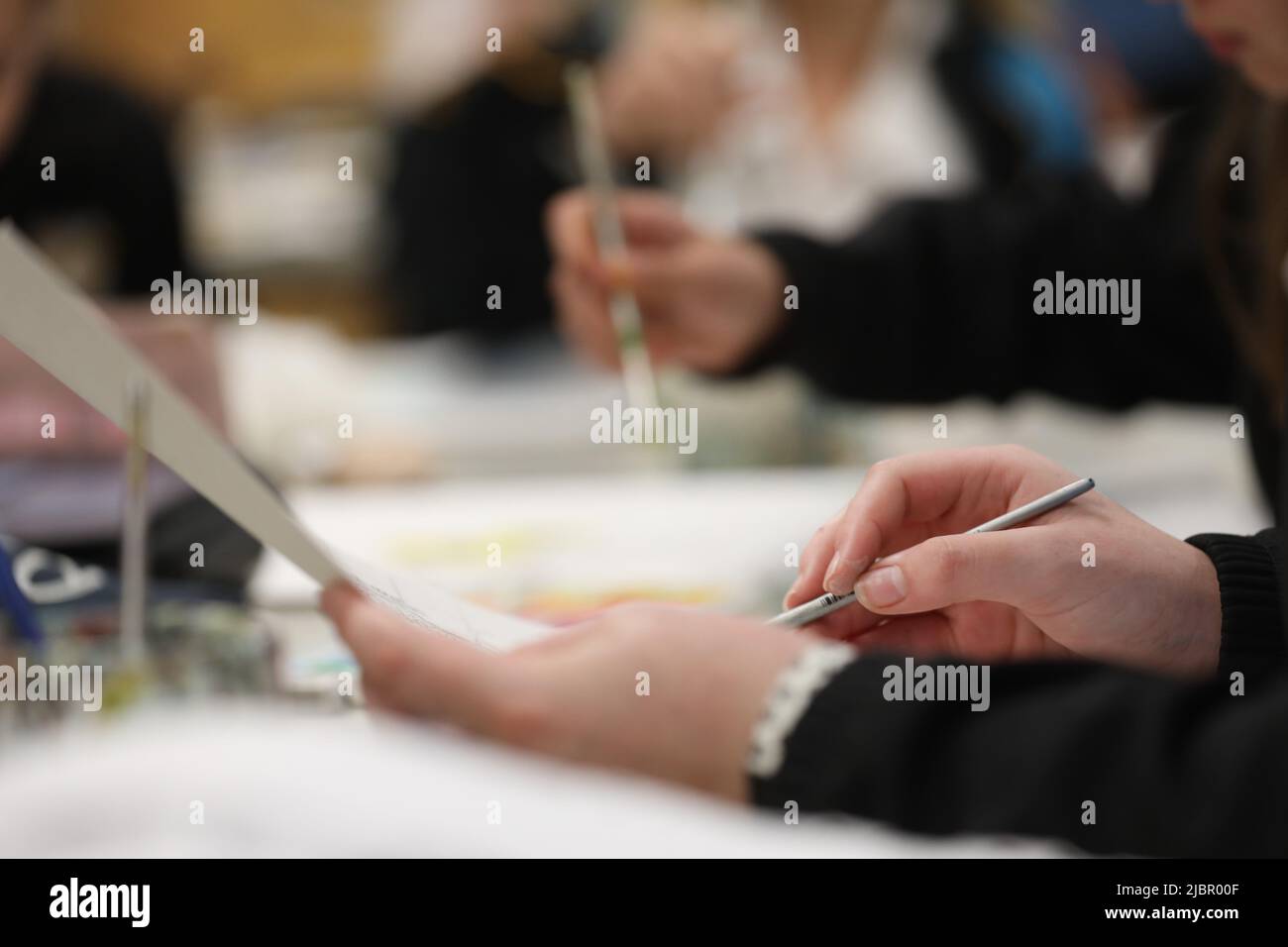 Eine Nahaufnahme einer Hand von Kunststudenten, die einen kleinen grauen Pinsel mit Wasserfarbe an einem Schreibtisch in einem Kunstschulzimmer hält, in dem andere Schüler malen Stockfoto