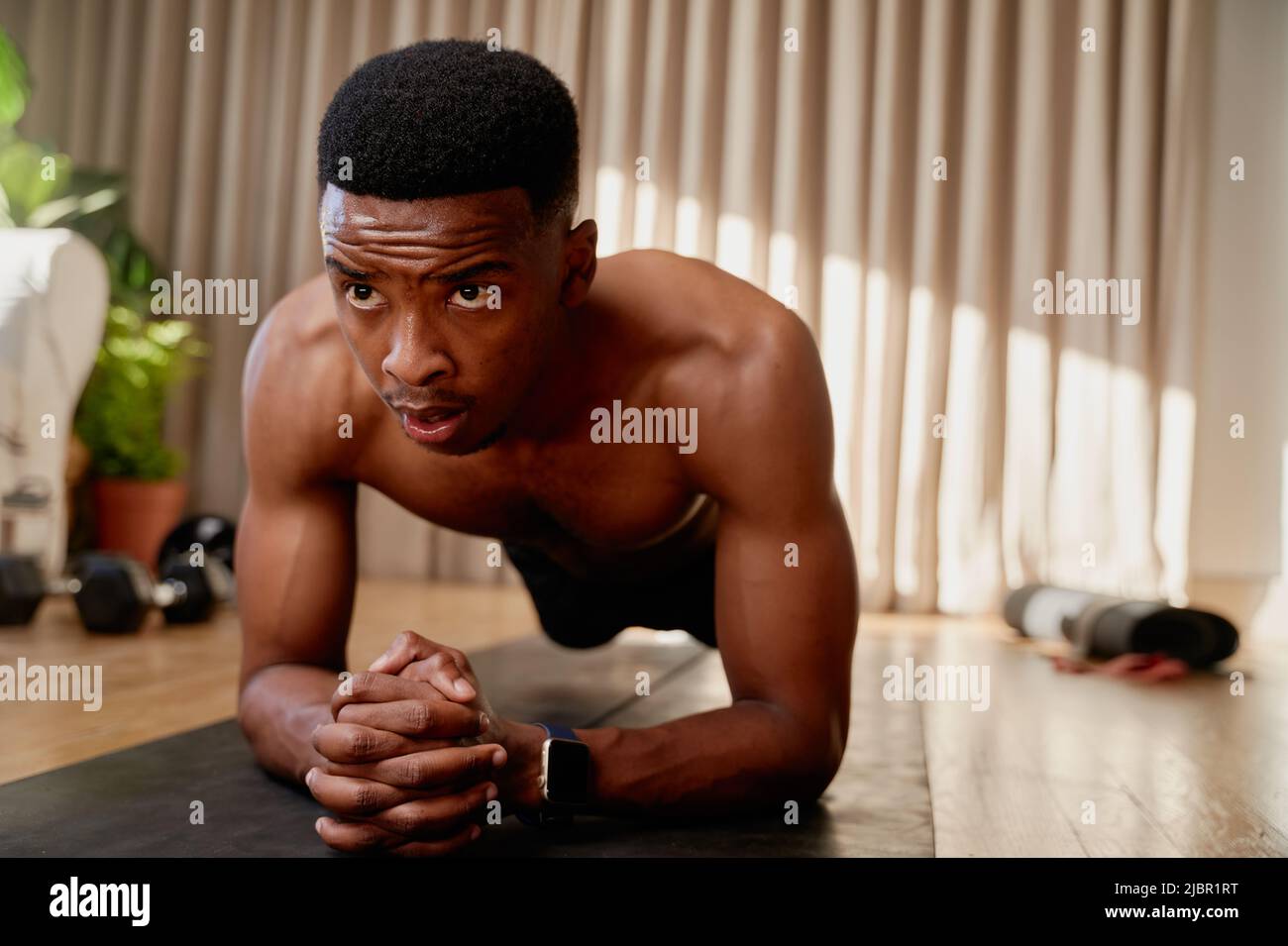 Junger afroamerikanischer schwarzer Mann, der zu Hause trainiert, hält eine Plankenposition und trainiert von zu Hause aus auf seiner Trainingsmatte Stockfoto