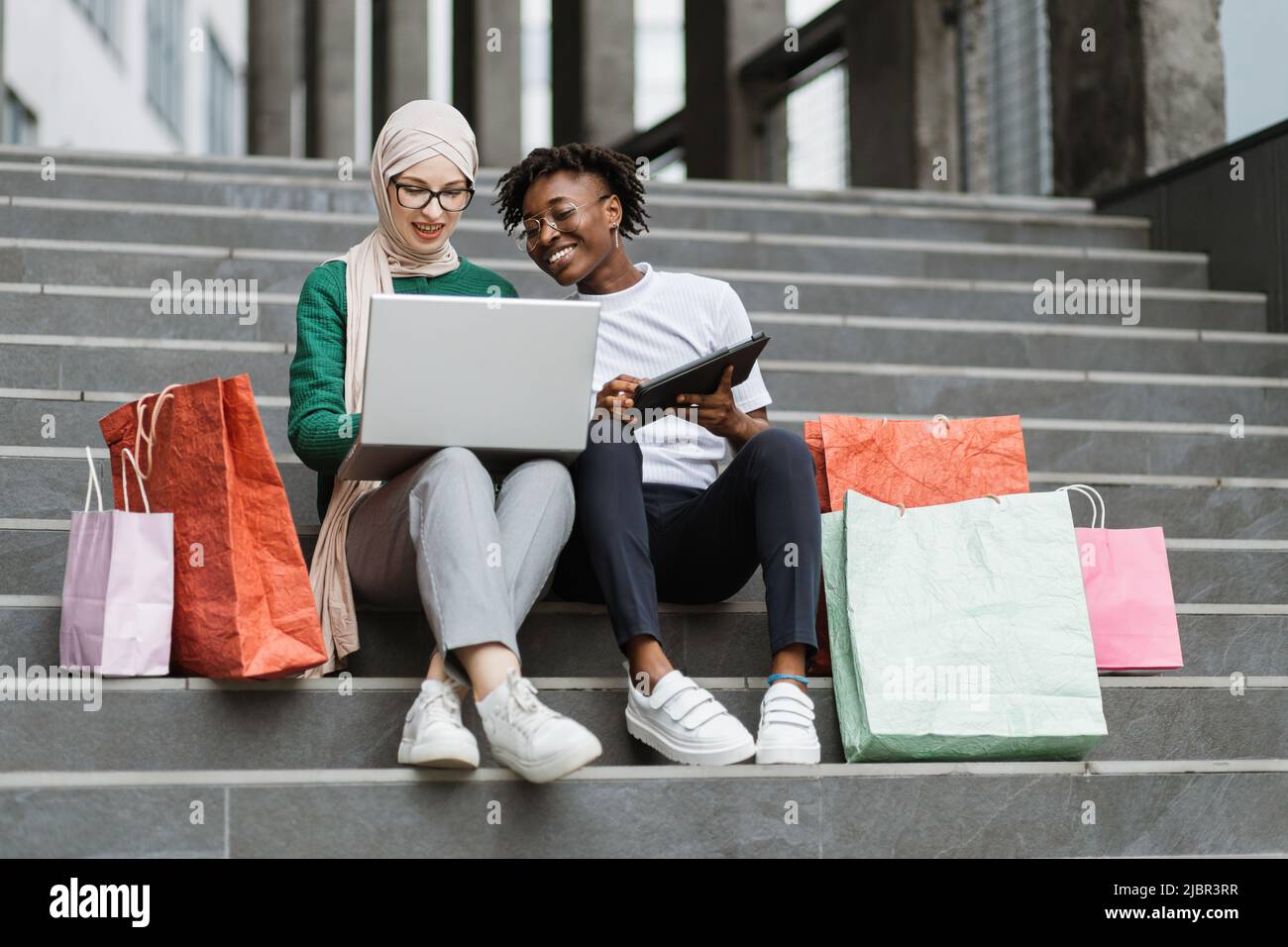 Outdoor in voller Länge Fashion Portrait von zwei jungen multirassischen  Damen in stilvoller Kleidung sitzen auf den Stufen eines großen  Einkaufszentrums und bestellen Kleidung online farbige Einkaufstaschen  liegen neben ihnen Stockfotografie -