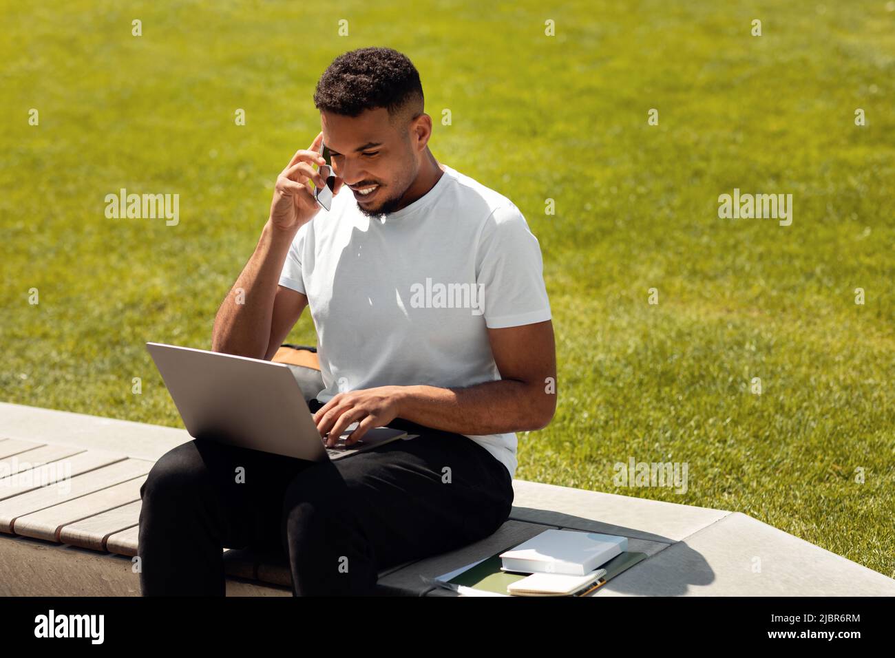 Positiver afroamerikanischer Mann, der im Freien in der Stadt sitzt, mit dem Handy spricht, während er einen Laptop benutzt und draußen arbeitet Stockfoto
