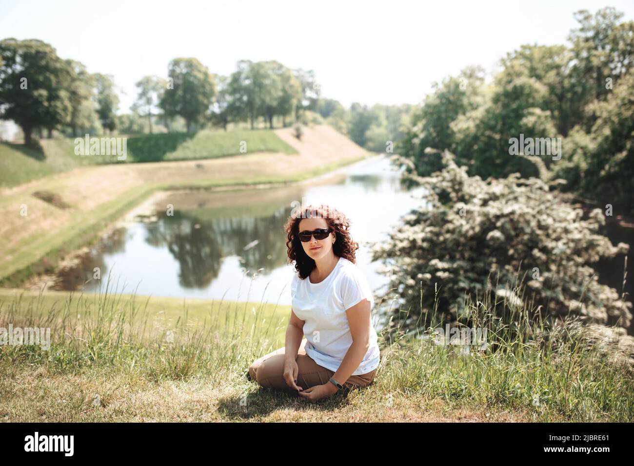 Die Reisende sitzt auf grünem Gras und sieht gerade aus. Die Frau genießt die Natur und sitzt am Ufer des Grabens rund um das Schloss Castellet. Festung aus dem 17. Jahrhundert mit Museum in Kopenhagen, Dänemark. Weichfokus Stockfoto