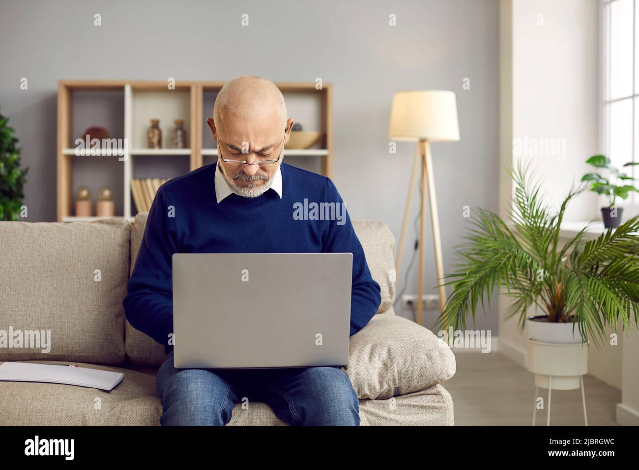 Älterer Mann in einer Brille sitzt zu Hause auf der Couch und arbeitet an seinem Laptop-Computer Stockfoto