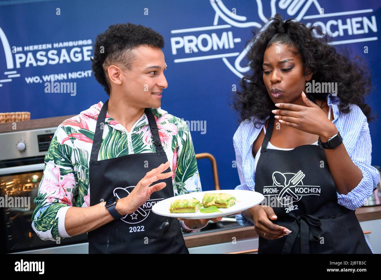 Hamburg, Deutschland. 08.. Juni 2022. Linda Nobat (r), Model, probiert die noch heiße Low Carb Lachsrolle von Prince Damien Ritzinger (l), Sängerin und Musical-Performerin. Um der Hamburger Lebensmittelbank in Not ein wenig zu helfen, findet in der Europa-Passage eine Wohltätigkeitsveranstaltung statt. Bis Samstag kochen dort Prominente mittags ihre Lieblingsgerichte für Besucher des Einkaufszentrums. Quelle: Jonas Walzberg/dpa/Alamy Live News Stockfoto