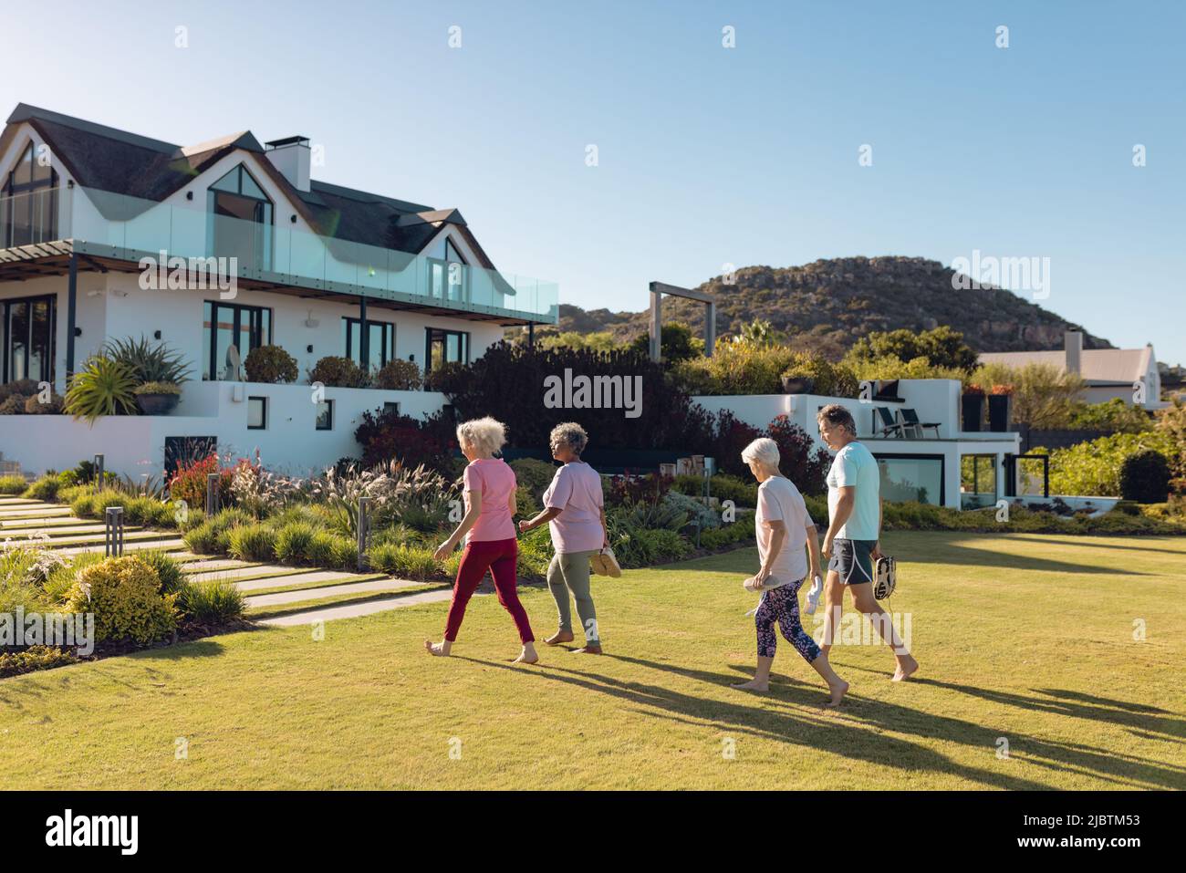 Mehrrassische Senioren, Männer und Frauen, die auf Gras in Richtung Pflegeheim gehen, gegen den klaren Himmel Stockfoto