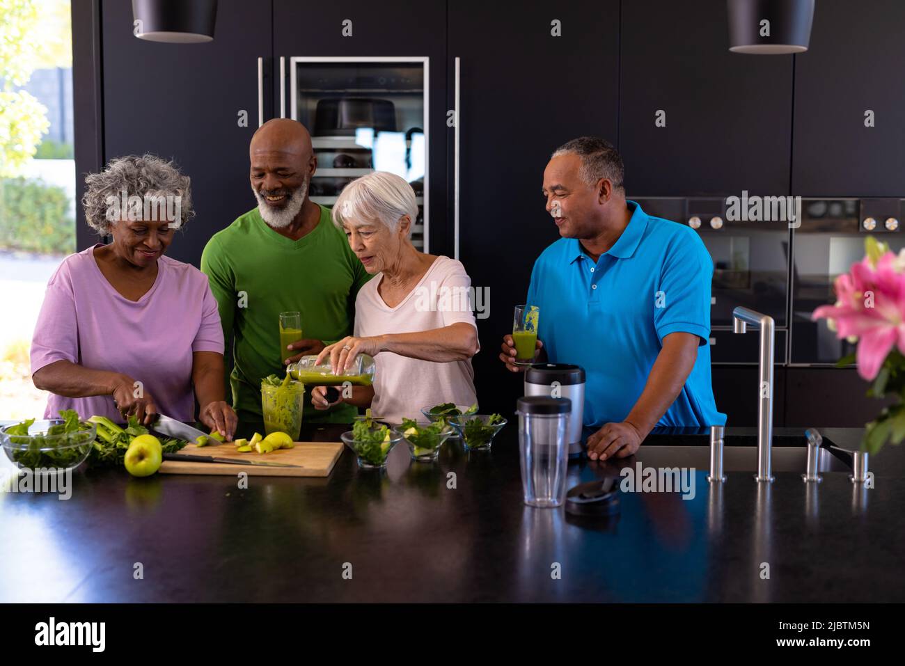 Mehrrassische ältere Männer, die Gläser mit Smoothie halten und Frauen betrachten, die sie in der Küche zubereiten Stockfoto