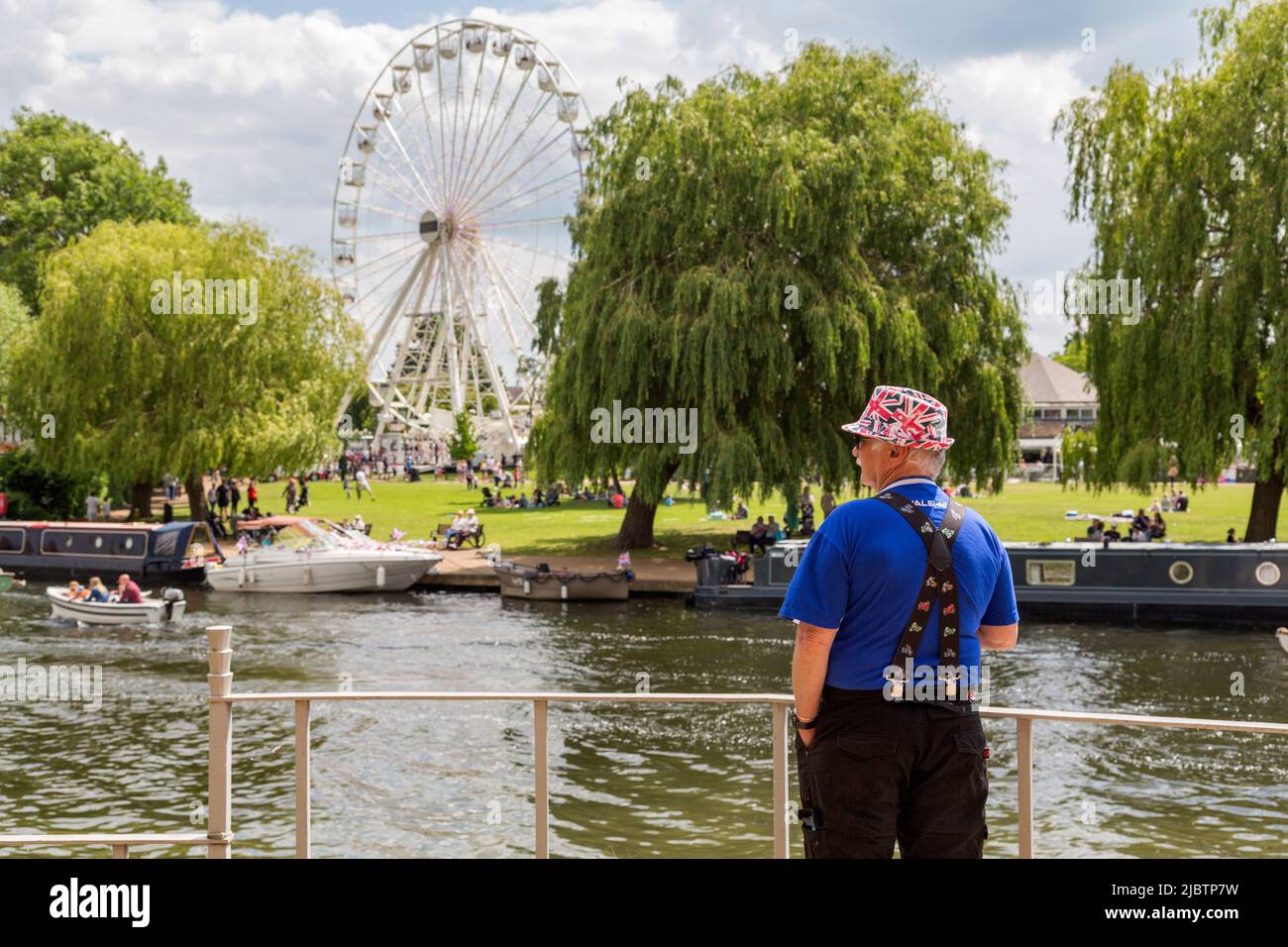 Ein Mann in einem Hut der Union Flag blickt über einen Fluss und sieht Menschen, die sich entspannen und die Sonne in einem Park genießen. Britisches Urlaubs-, Tourismus- oder Reisekonzept. Stockfoto