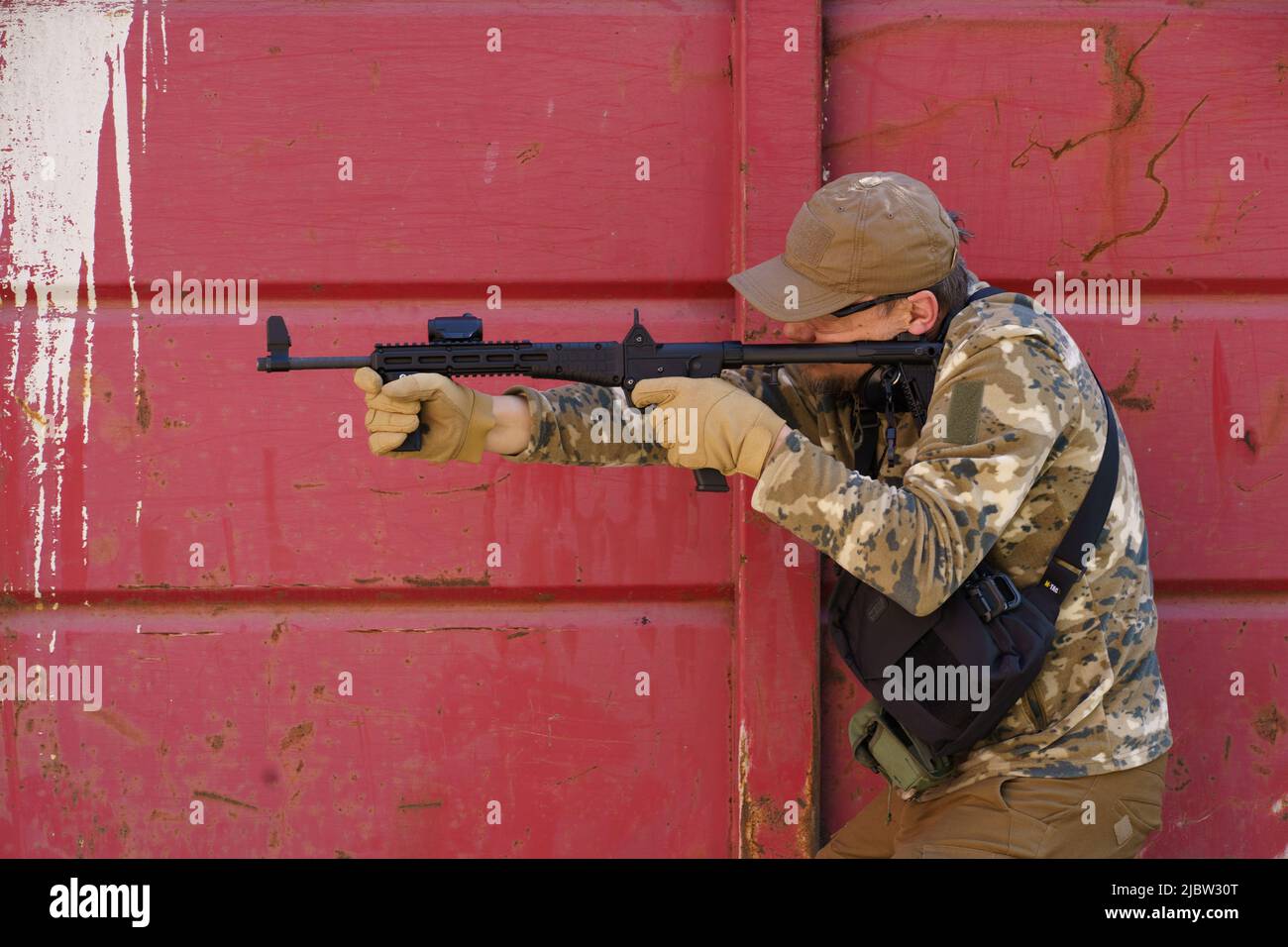 Militärsoldat Mann im Boot-Lager. Mann in taktischer Uniform mit Waffe oder Waffe. Taktisches Kampftraining im Lager. Schütze Mann mit Waffe in Militäruniform verstecken sich hinter rostigen roten Wand. Maskuline Hobbytätigkeit. Stockfoto