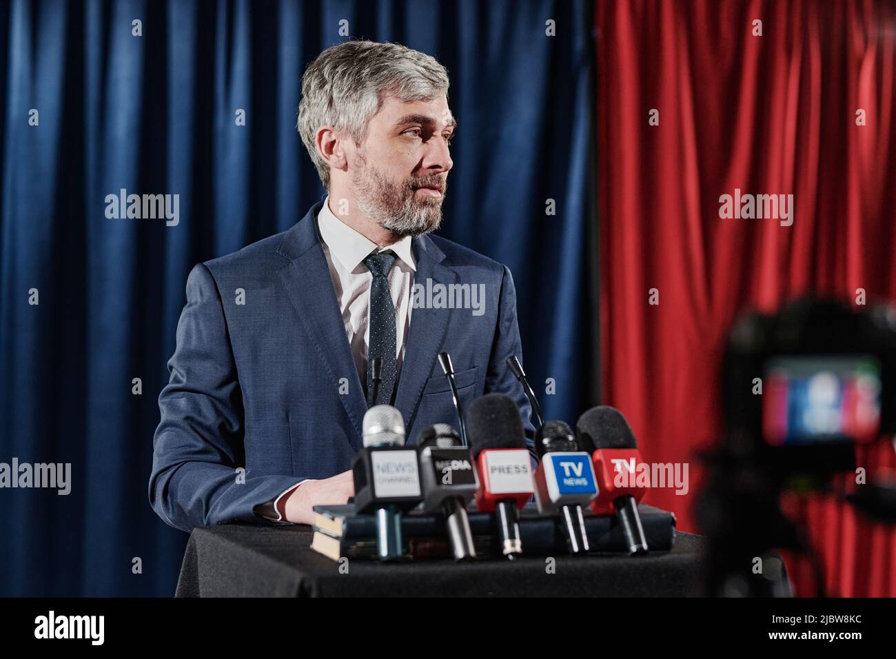 Reifer bärtiger Geschäftsmann, der mit Mikrofonen auf der Bühne steht und während der Online-Übertragung auf der Pressekonferenz ein Interview gibt Stockfoto