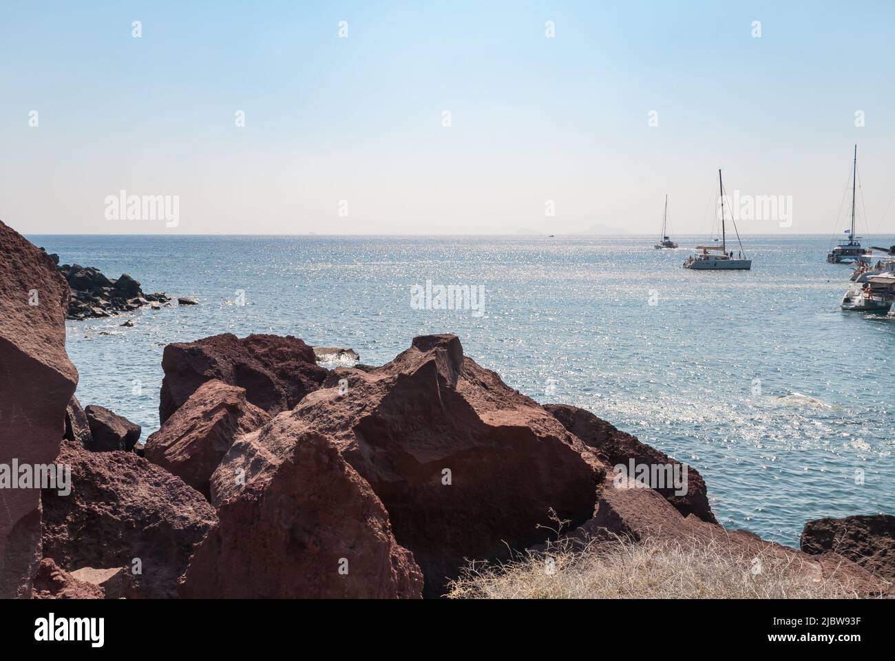 Rote Felsen an einem Inselstrand und Blick auf Segelboote und den Horizont weit voraus Stockfoto