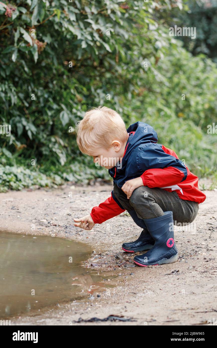 Ein Kind in einer roten Jacke und Gummistiefeln erkundet den Schlamm eines Sumpfes in der Natur bei einer Wanderung an der frischen Luft. Stockfoto