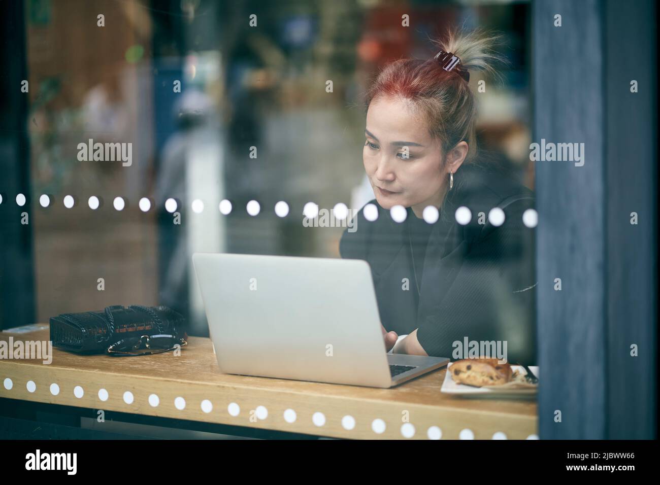 Durch eine Glasaufnahme einer asiatischen Geschäftsfrau mit mittlerem Erwachsenen, die im Café mit einem Laptop arbeitet Stockfoto