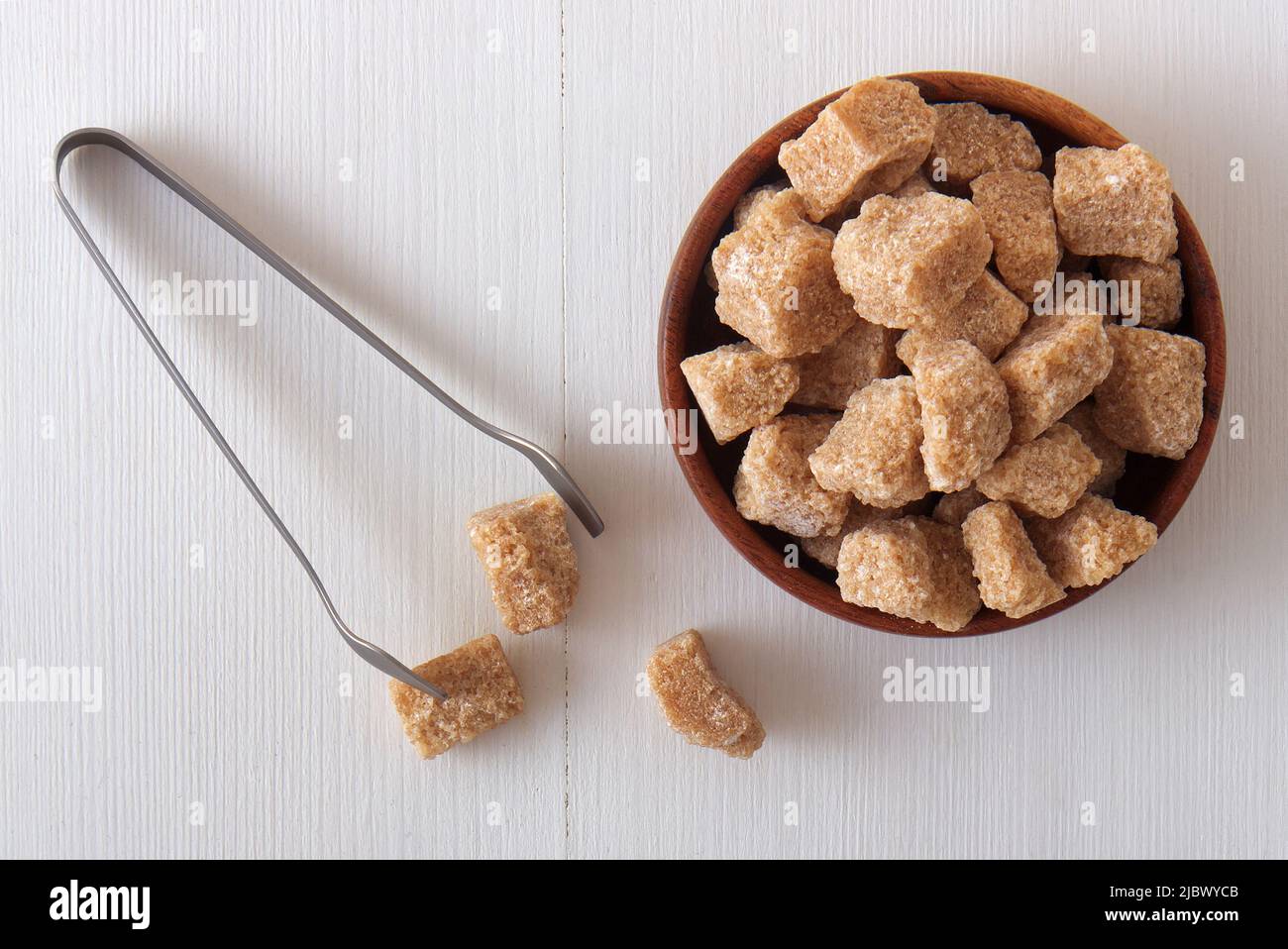 Würfelförmiger Rohrzucker in Holzschüssel auf einem weißen Holztisch mit silberner Zuckerzange. Nahaufnahme aus hoher Perspektive, keine Menschen. Stockfoto