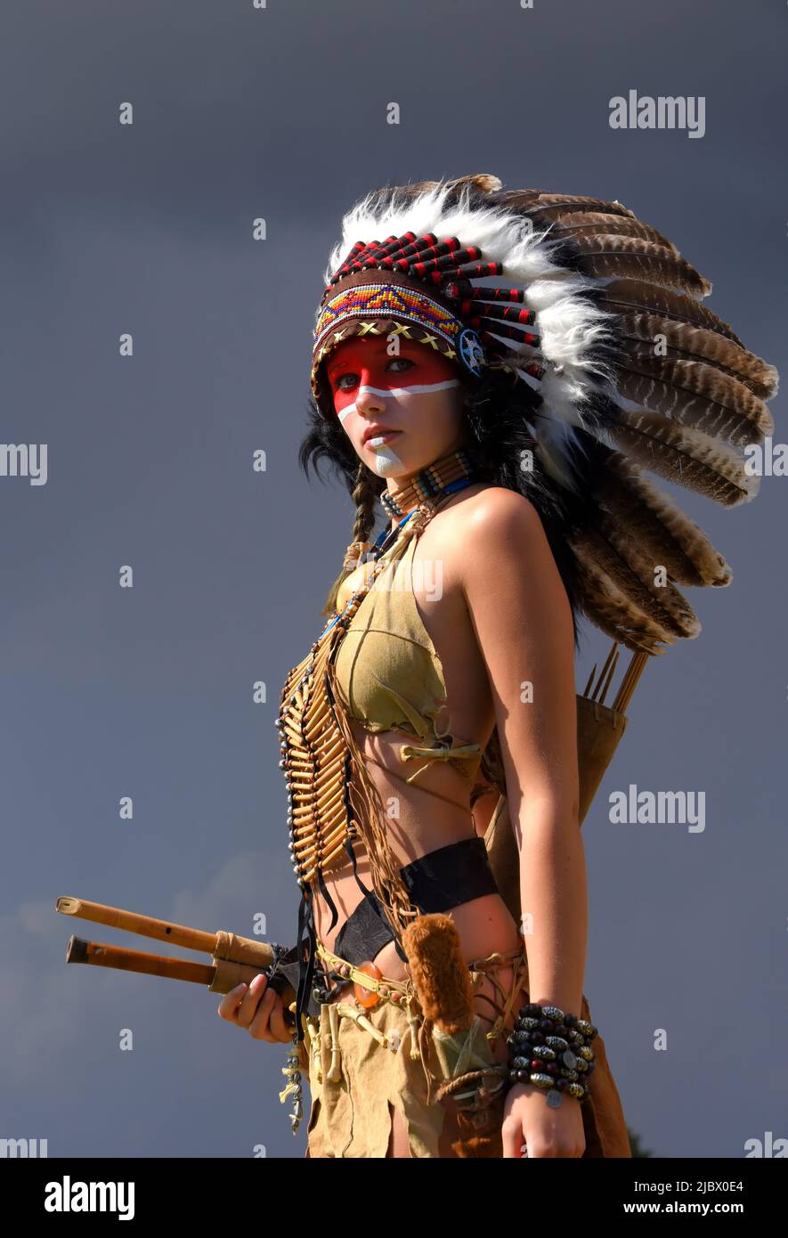 Eine indianische Ureinwohnerin steht vor grauen Regenwolken. Sie trägt einen gefiederten Kopfschmuck und trägt traditionelle indische Kleidung. Stockfoto