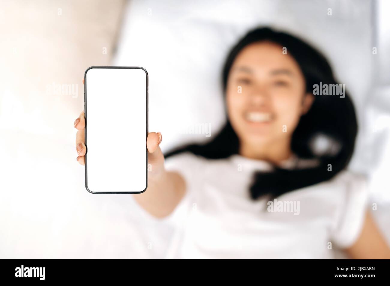 Mock-up-Konzept für Werbung oder Präsentation. Defokussierte asiatische Mädchen liegt in einem Bett, lächelnd, zeigt ein Smartphone mit leeren weißen Bildschirm, Kopierraum. Wireless-Technologien Stockfoto