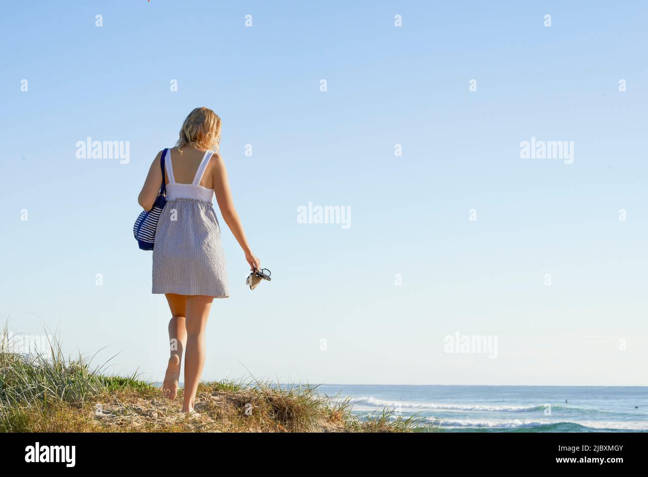Rückansicht einer jungen Frau, die ihre Tasche und Schuhe trägt und am Strand entlang einer Sandküste läuft Stockfoto