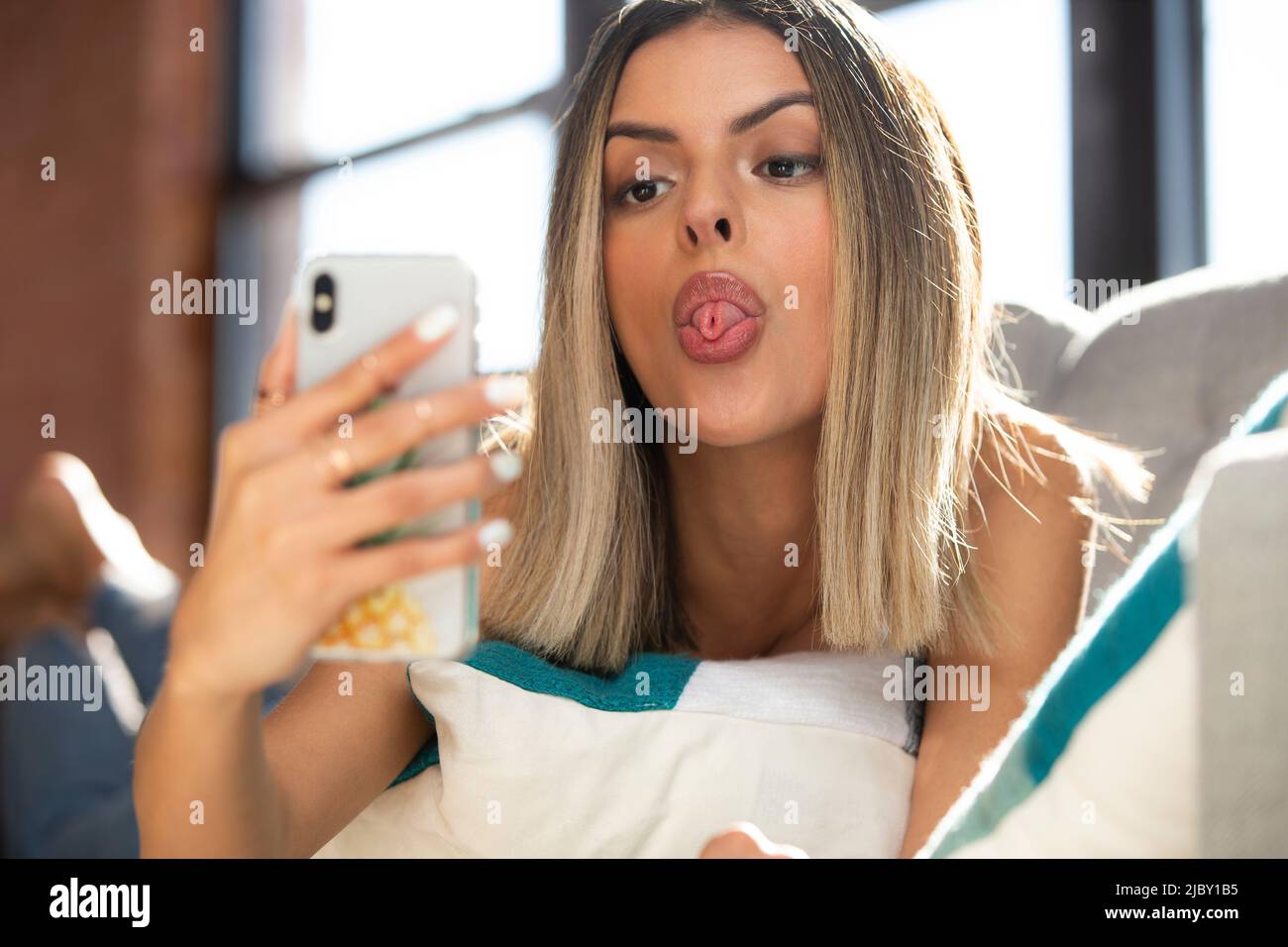 ait der hispanischen Frau, die ein weißes Tanktop und eine zerrissene blaue Jeans trägt, auf der grauen Couch faulenzt und mit dem Mobiltelefon ein Selfie macht, während sie aus der Zunge ragt. Stockfoto