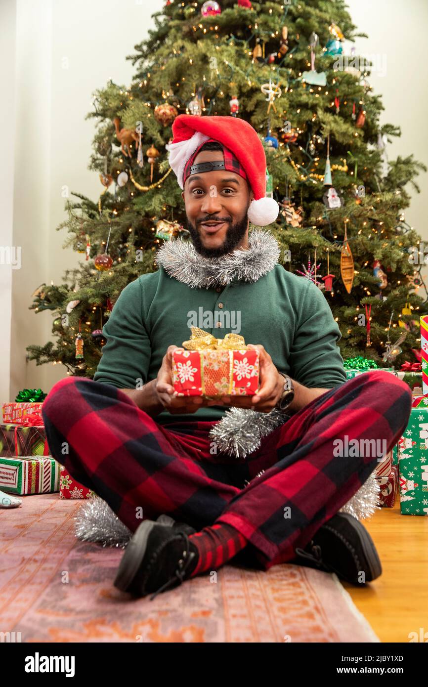 Porträt eines Mannes mit Weihnachtsgeschenk, der vor einem Baum sitzt und einen Weihnachtsmütze trägt. Stockfoto