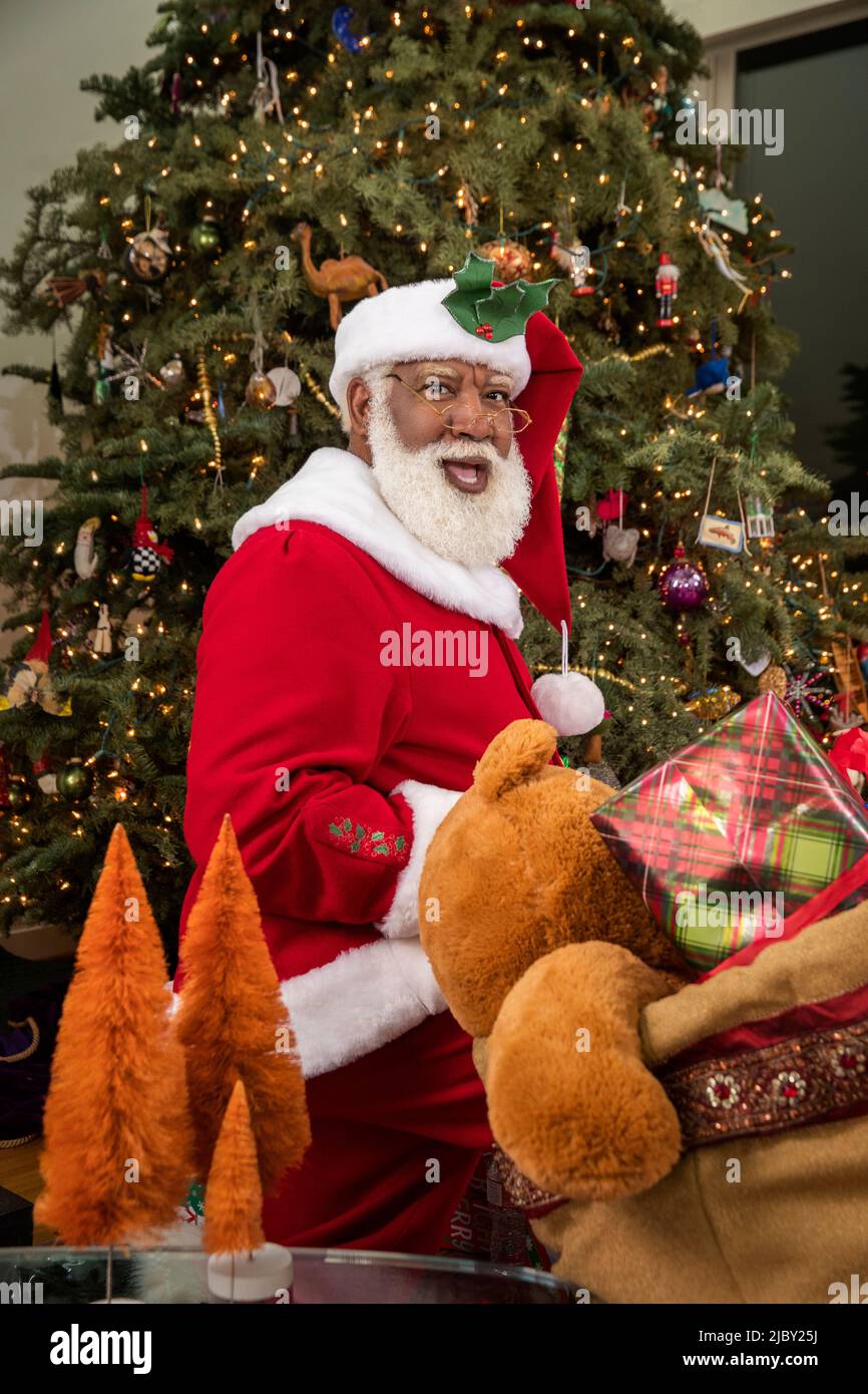 Ein überraschender Weihnachtsmann blickt über seine Schulter auf die Kamera und platziert Geschenke unter dem Weihnachtsbaum. Serie „Black Santa“. Stockfoto