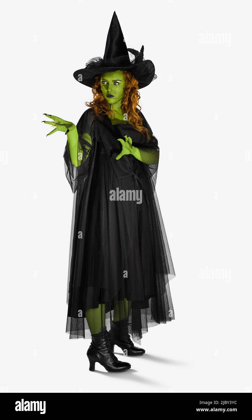 Porträt einer Frau, die als Hexe gekleidet ist, vor einem weißen Hintergrund mit grüner Körperfarbe, die mit den Händen nach oben aus der Kamera schaut und einen Zauber auswirft. Stockfoto