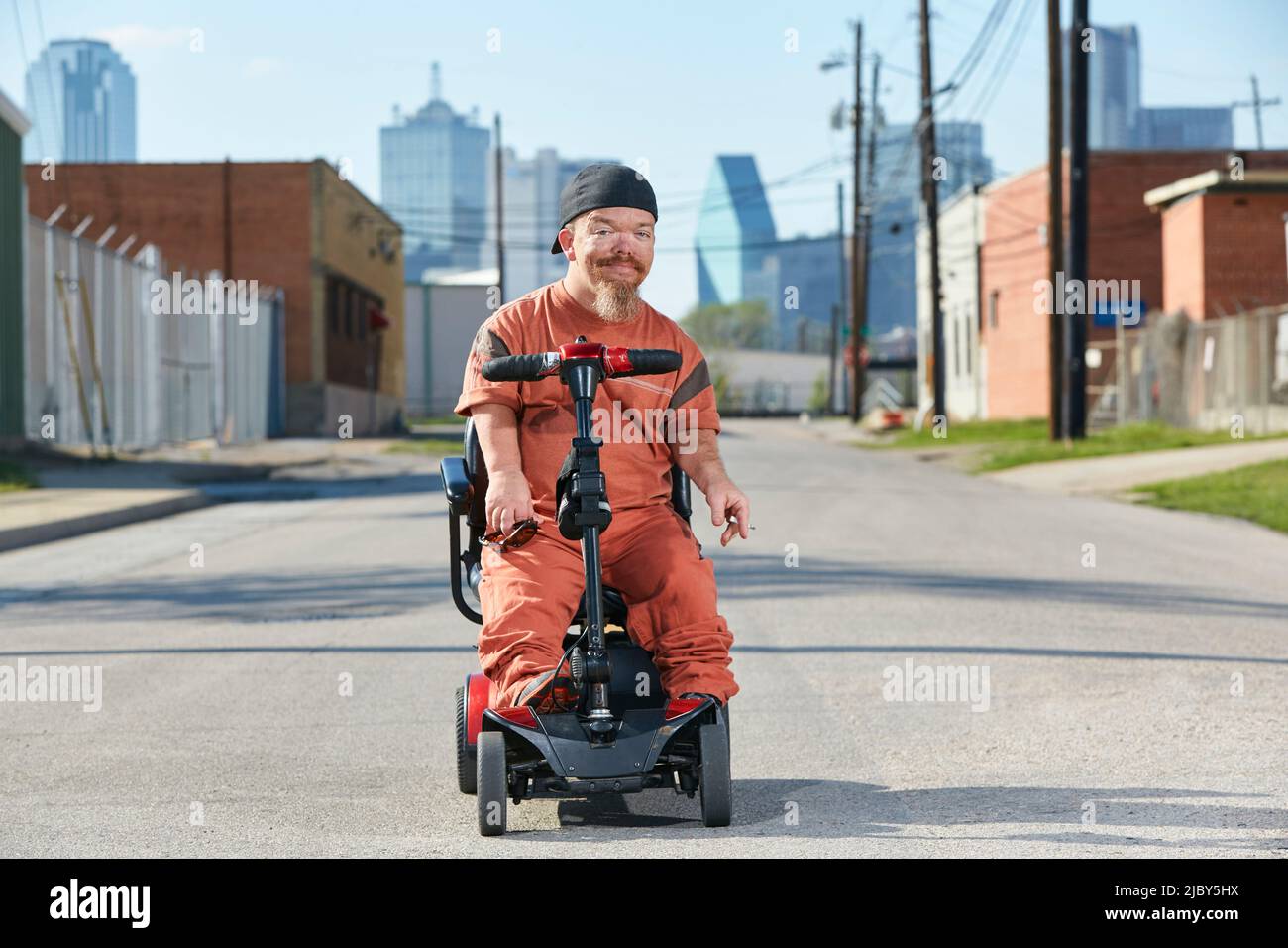 Porträt eines männlichen erwachsenen kleinen Menschen auf der Straße in Texas auf einem Mobilitätsroller, der in die Kamera blickt, mit der Skyline von Dallas im Hintergrund. Stockfoto