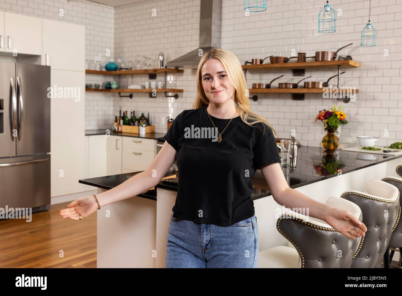 Porträt einer jungen Frau, die in ihrer Küche steht und ihre Arme mit einem einladenden Ausdruck in die Kamera blickt. Stockfoto