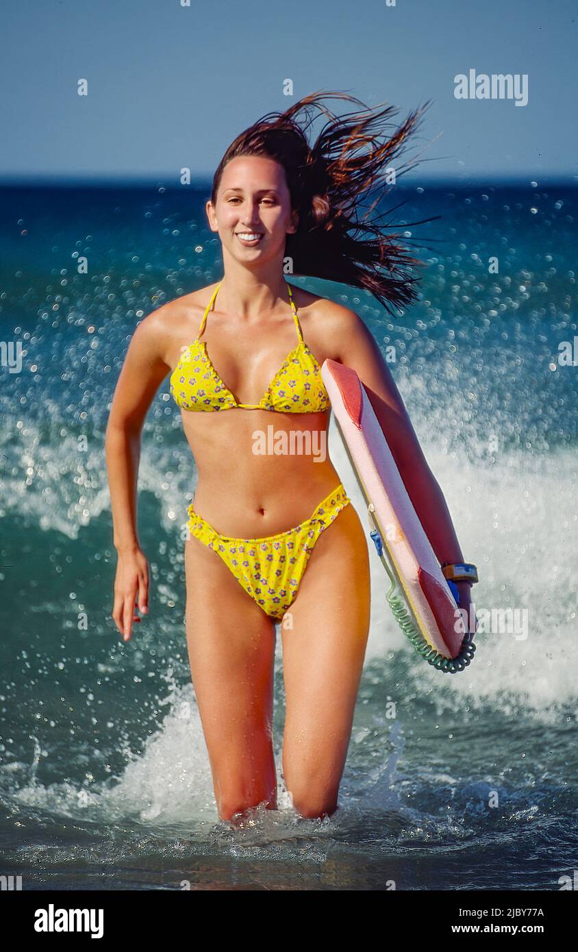 Junge Frau, die am Strand mit ihrem Boggie Board durch das Wasser läuft und lächelt Stockfoto