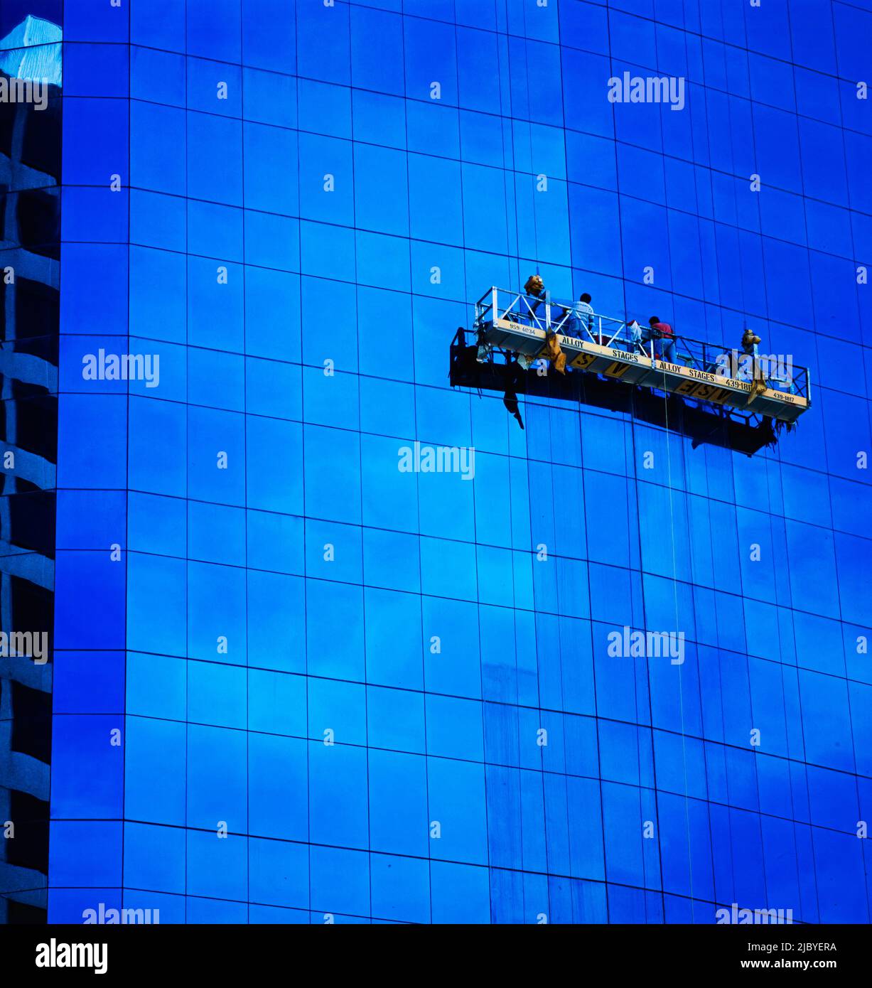 Zwei Männer in hängenden Gerüsten, die die Fenster eines Hochhauses aus Glas putzen und Wolken in Glas spiegeln Stockfoto