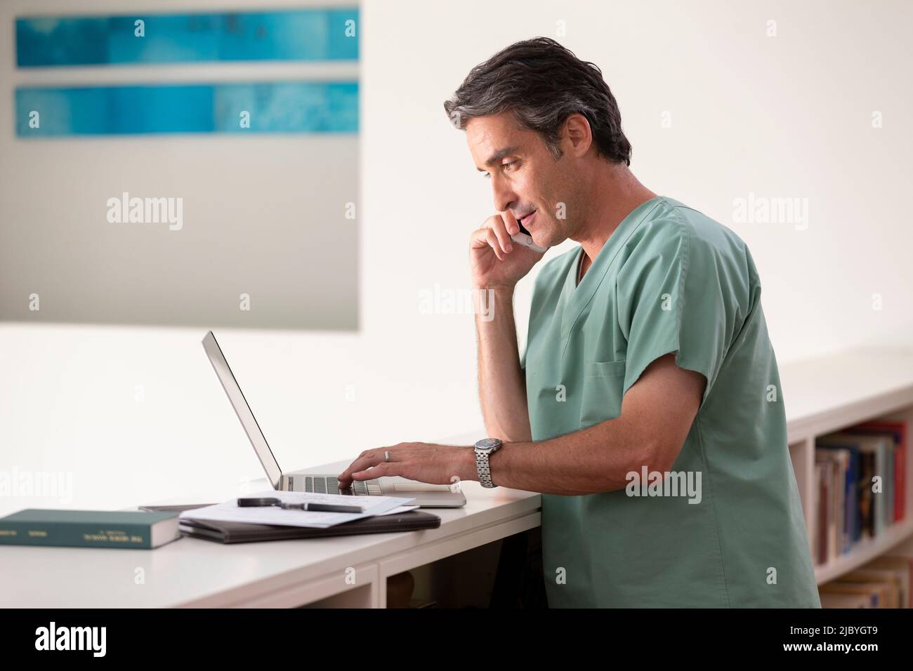 Hispanischer Arzt in seinem Haus, der Telemedizin praktiziert, mit dem Handy telefoniert und einen Laptop benutzt Stockfoto