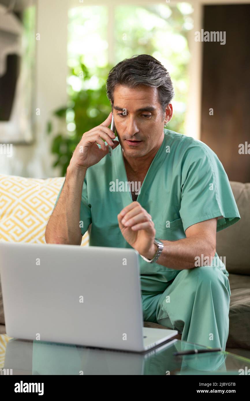 Hispanischer Arzt, der von zu Hause aus Telemedizin praktiziert, Handy und Laptop benutzt und mit dem Patienten während eines Videoanrufs spricht Stockfoto