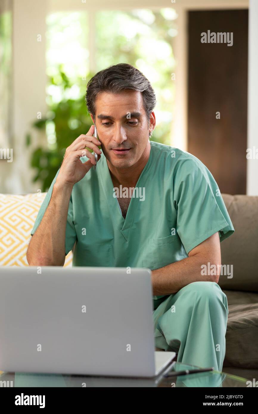 Hispanischer Arzt, der von zu Hause aus Telemedizin praktiziert, Handy und Laptop benutzt und mit dem Patienten während eines Videoanrufs spricht Stockfoto