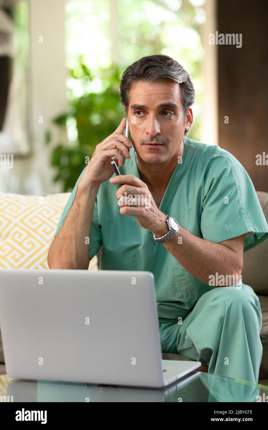 Hispanischer Arzt, der von zu Hause aus Telemedizin praktiziert, Handy und Laptop benutzt und dem Patienten bei einem Videoanruf zugehört Stockfoto
