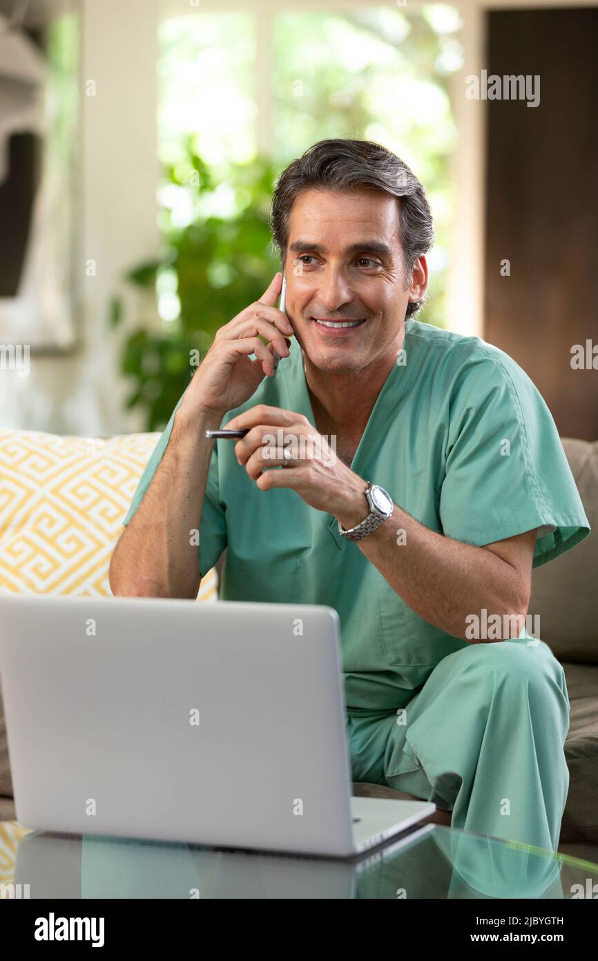 Hispanischer Arzt, der von zu Hause aus Telemedizin praktiziert, Handy und Laptop benutzt und dem Patienten bei einem Videoanruf zugehört Stockfoto