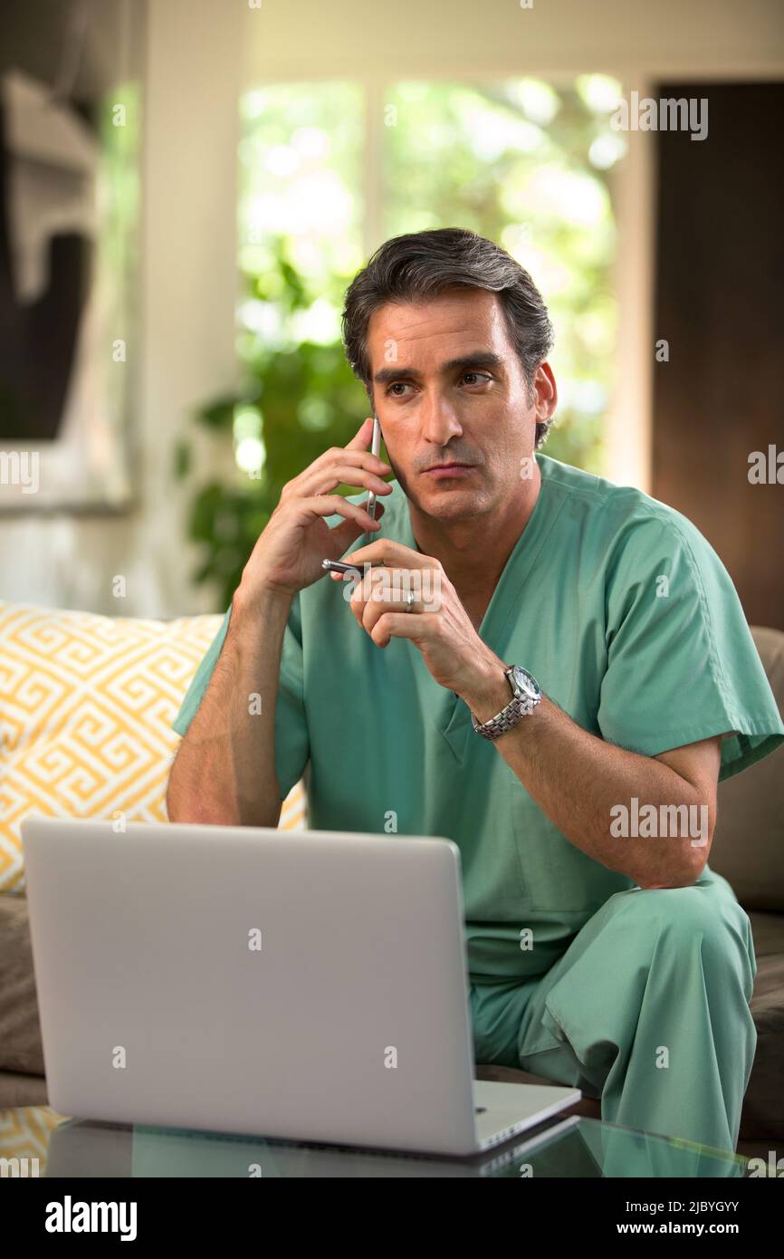 Hispanischer Arzt, der von zu Hause aus Telemedizin praktiziert, Handy und Laptop benutzt und dem Patienten bei einem Videoanruf zugehört Stockfoto