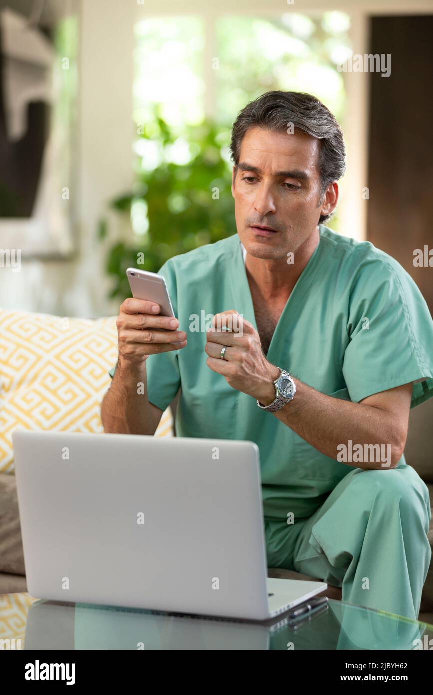 Hispanischer Arzt, der von zu Hause aus Telemedizin praktiziert, Handy und Laptop benutzt und dem Patienten bei einem Videoanruf zugehört Stockfoto