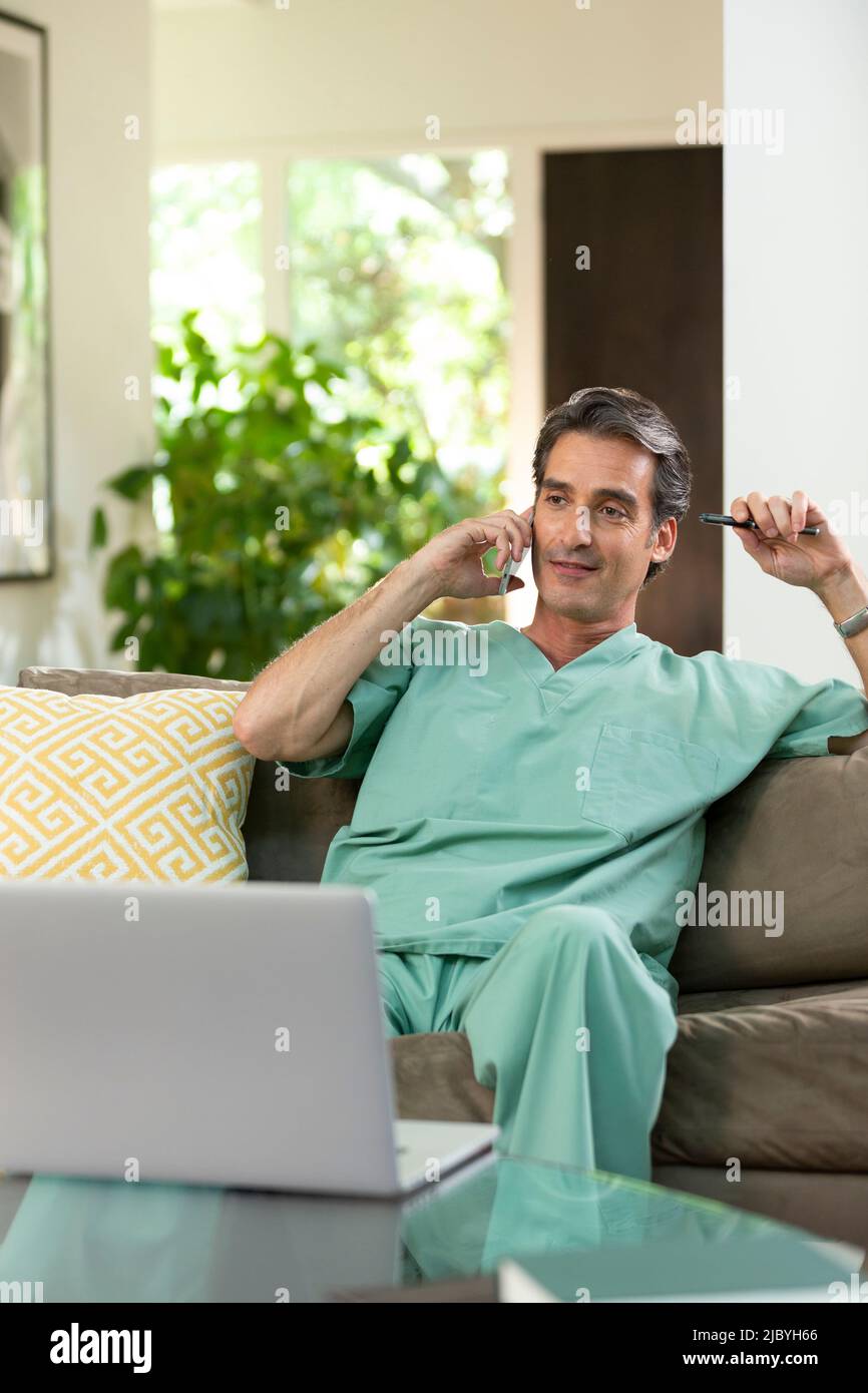 Hispanischer Arzt, der von zu Hause aus Telemedizin praktiziert, Handy und Laptop benutzt und dem Patienten bei einem Videoanruf zugehört Stockfoto