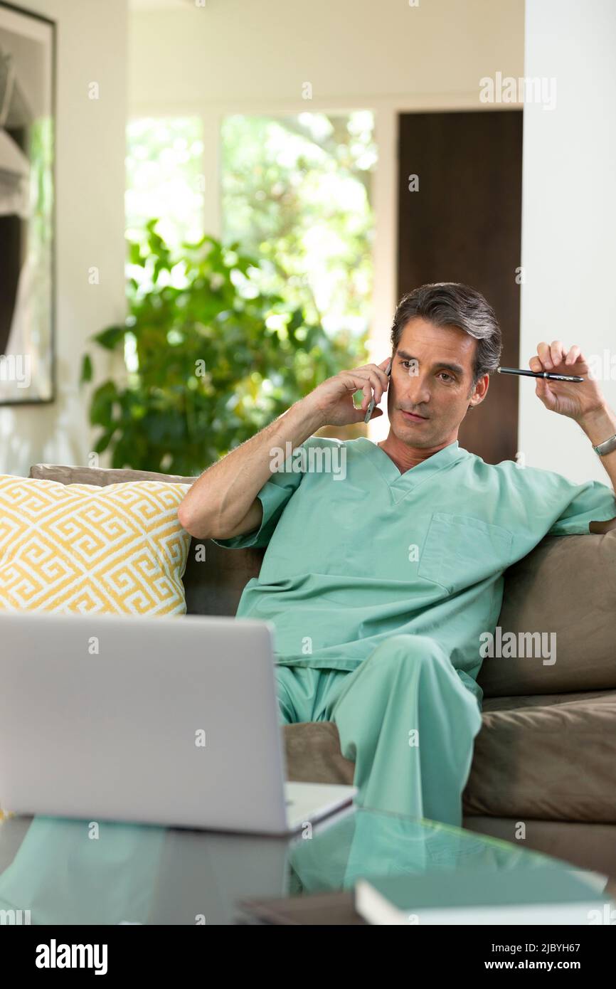 Hispanischer Arzt, der von zu Hause aus Telemedizin praktiziert, Handy und Laptop benutzt und dem Patienten bei einem Videoanruf zugehört Stockfoto