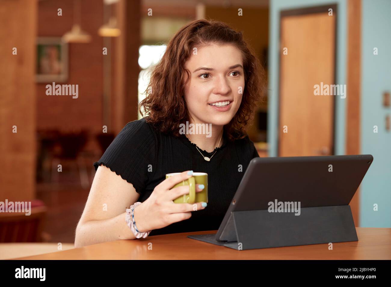 Porträt einer jungen kaukasischen Frau, die schwarzes T-Shirt trägt, an der Bar in der Küche des Loft in der Innenstadt mit einem Kaffeebecher mit Tablet und Tastatur sitzt Stockfoto