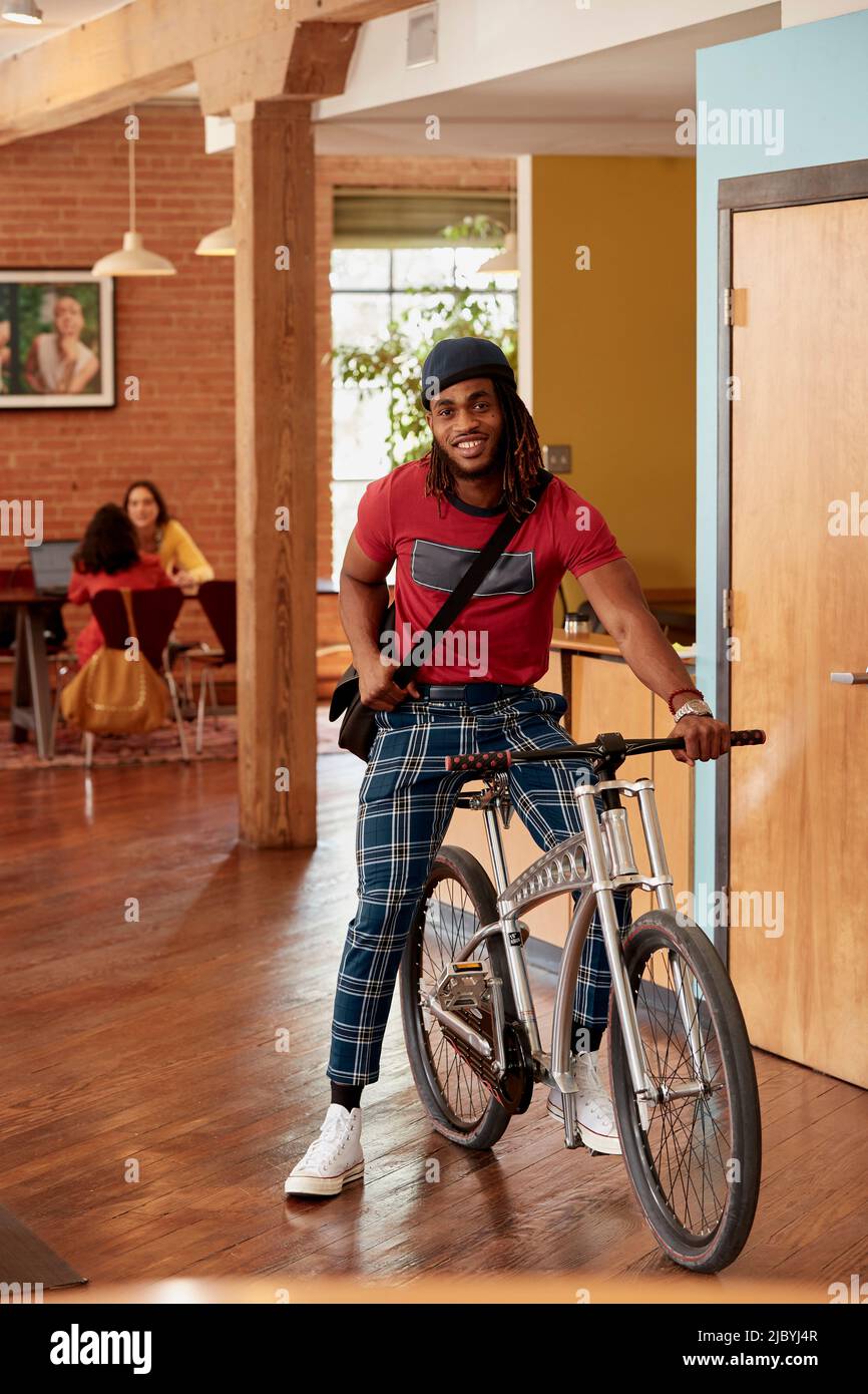 Porträt eines jungen ethnischen Mannes mit karierten Hosen und Strickmütze, der mit dem Fahrrad im öffentlichen Bereich des Bürolofts in der Innenstadt steht Stockfoto